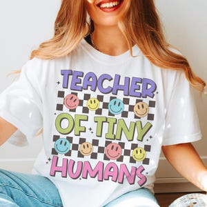 Peut inclure: T-shirt blanc avec le texte "Teacher of Tiny Humans" en lettres color&eacute;es et un motif &agrave; carreaux avec des visages souriants.