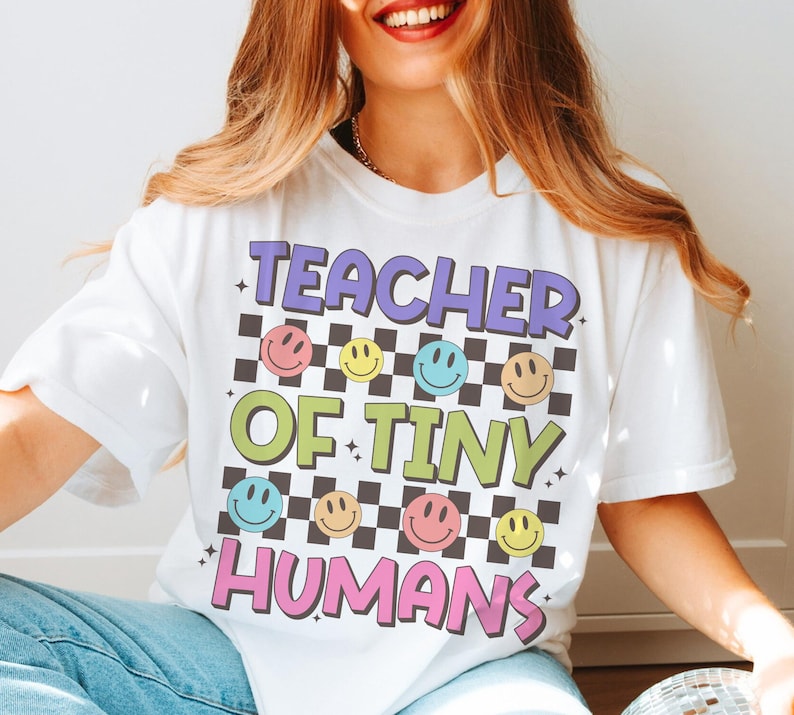 Peut inclure: T-shirt blanc avec le texte "Teacher of Tiny Humans" en lettres color&eacute;es et un motif &agrave; carreaux avec des visages souriants.