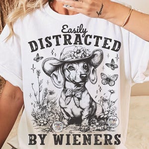 May include: White t-shirt with a black and white illustration of a dachshund wearing a flower crown and the text "Easily Distracted By Wieners".