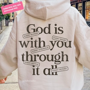 May include: Cream-colored hoodie with the text "God is with you through it all" in dark brown. The text is surrounded by smaller phrases like "Every Season" and "Every Doubt". The hoodie has a hood and long sleeves.
