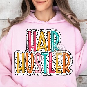 May include: Pink hoodie with a white and black polka dot background and the text "HAIR HUSTLER" in colorful letters.