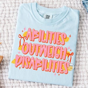 May include: Light blue t-shirt with the text "ABILITIES OUTWEIGH DISABILITIES" in pink and orange lettering. The shirt has a crew neck and short sleeves. The text is decorated with ribbons and stars.