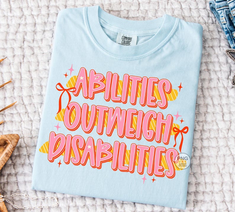 May include: Light blue t-shirt with the text "ABILITIES OUTWEIGH DISABILITIES" in pink and orange lettering. The shirt has a crew neck and short sleeves. The text is decorated with ribbons and stars.