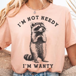 May include: A peach-coloured t-shirt with a black and white illustration of a raccoon. The text "I'm not needy I'm wanty" is printed on the shirt.