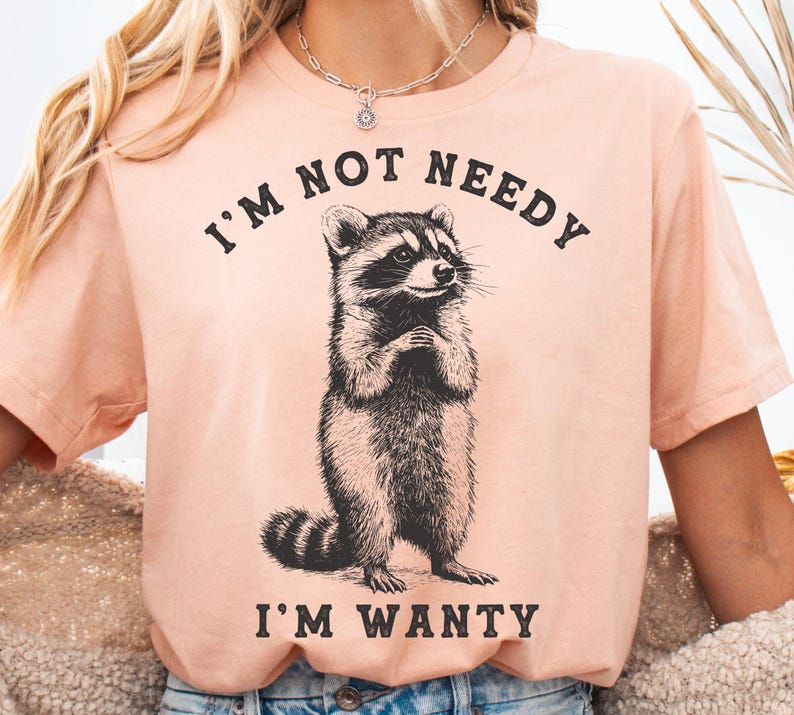 May include: A peach-coloured t-shirt with a black and white illustration of a raccoon. The text "I'm not needy I'm wanty" is printed on the shirt.