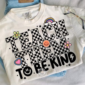 以下が含まれることがあります： クリーム色のTシャツ。 「TEACH THEM TO BE KIND」の文字がチェッカーボード柄でプリントされています。虹、花、星、ハート、スマイリーフェイスが特徴です。丸いメガネがTシャツの上に置かれています。