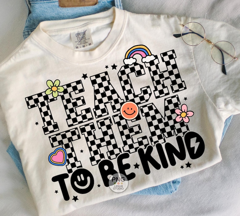 以下が含まれることがあります： クリーム色のTシャツ。 「TEACH THEM TO BE KIND」の文字がチェッカーボード柄でプリントされています。虹、花、星、ハート、スマイリーフェイスが特徴です。丸いメガネがTシャツの上に置かれています。