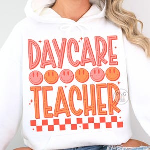 Peut inclure: Sweat-shirt blanc avec le texte "DAYCARE TEACHER" en lettres roses et orange. Le texte est entour&eacute; d'une rang&eacute;e de six visages souriants roses et orange et d'un motif &agrave; carreaux rouge et blanc en bas.