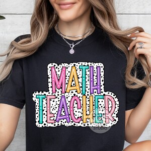 Op de afbeelding: Zwart T-shirt met een kleurrijke afbeelding waarop "MATH TEACHER" staat in een vetgedrukte, omrande lettertype. De afbeelding is omgeven door witte stippen.