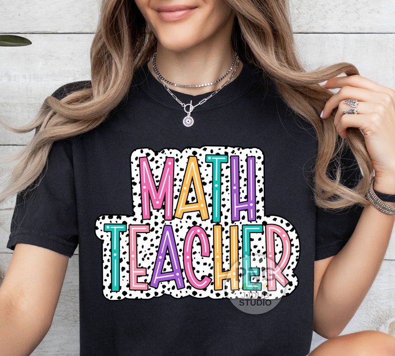 Op de afbeelding: Zwart T-shirt met een kleurrijke afbeelding waarop "MATH TEACHER" staat in een vetgedrukte, omrande lettertype. De afbeelding is omgeven door witte stippen.