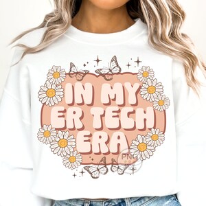 May include: White sweatshirt with a pink and brown graphic that says "In My ER Tech Era" surrounded by daisies and butterflies.