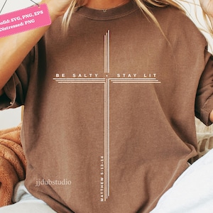 以下が含まれることがあります： 白い十字のデザインが施された茶色のTシャツ。十字は中央に配置され、「BE SALTY STAY LIT」と「MATTHEW 5:13-14」の文字が書かれています。 ゆったりとしたフィット感で、柔らかい素材で作られています。