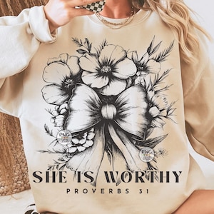 Peut inclure: Un sweat-shirt beige avec un motif floral noir et blanc et un n&oelig;ud. Le texte "She is Worthy Proverbs 31" est imprim&eacute; en bas du sweat-shirt.