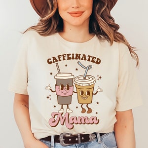 May include: A beige t-shirt with the text "caffeinated mama" in brown and pink lettering. The text is surrounded by two cartoon coffee cups with faces and arms.