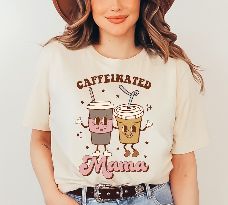 May include: A beige t-shirt with the text "caffeinated mama" in brown and pink lettering. The text is surrounded by two cartoon coffee cups with faces and arms.