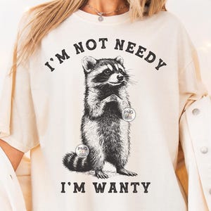 K&ouml;nnte beinhalten: Ein wei&szlig;es T-Shirt mit einer schwarz-wei&szlig;en Illustration eines Waschb&auml;ren. Der Text "I'm not needy I'm wanty" ist auf das T-Shirt gedruckt.