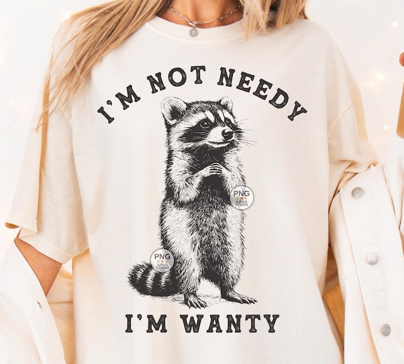 K&ouml;nnte beinhalten: Ein wei&szlig;es T-Shirt mit einer schwarz-wei&szlig;en Illustration eines Waschb&auml;ren. Der Text "I'm not needy I'm wanty" ist auf das T-Shirt gedruckt.