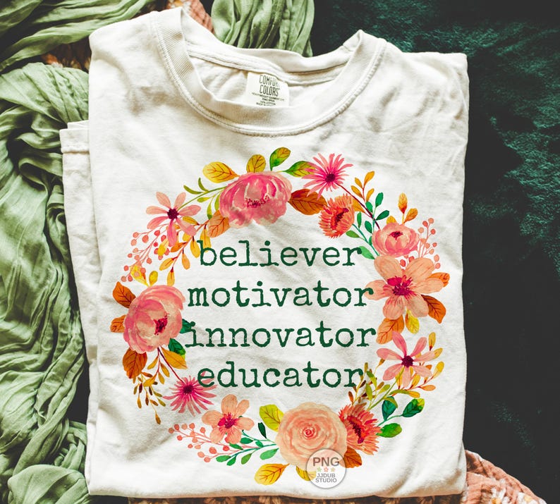 以下が含まれることがあります： 「believer, motivator, innovator, educator」の緑色の文字と花のリースデザインが特徴のクリーム色のTシャツ。 リースにはピンクとオレンジの花と緑の葉が含まれています。 シャツは折りたたまれています。