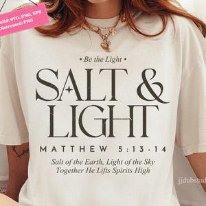 May include: Cream-colored t-shirt with the text "SALT & LIGHT" in a bold, dark font. Below the main text are the words "MATTHEW 5:13-14" and a quote. The shirt has a relaxed fit and short sleeves.