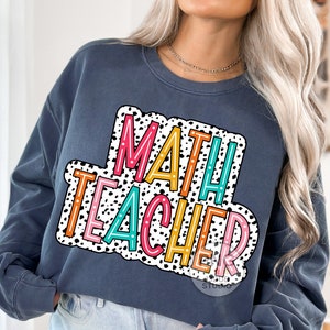 Op de afbeelding: Een marineblauw sweatshirt met een kleurrijke afbeelding waarop "MATH TEACHER" staat in een wit en zwart stippenpatroon.