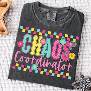 以下が含まれることがあります： カラフルで遊び心のあるデザインの「CHAOS Coordinator」という文字が入った濃いグレーのTシャツ。テキストはピンク、黄色、ターコイズの文字に花のアクセントが施されています。Tシャツはクルーネックで半袖です。