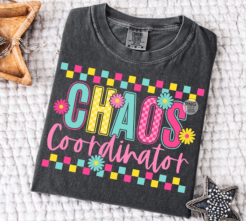 以下が含まれることがあります： カラフルで遊び心のあるデザインの「CHAOS Coordinator」という文字が入った濃いグレーのTシャツ。テキストはピンク、黄色、ターコイズの文字に花のアクセントが施されています。Tシャツはクルーネックで半袖です。