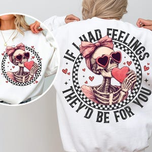 May include: White sweatshirt with a graphic of a skeleton wearing sunglasses and a pink bow holding a red heart. The text "If I had feelings they'd be for you" is printed around the graphic.