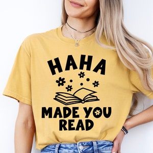 May include: A yellow t-shirt with black text that reads "HAHA MADE YOU READ" and a black illustration of a book with flowers above it.