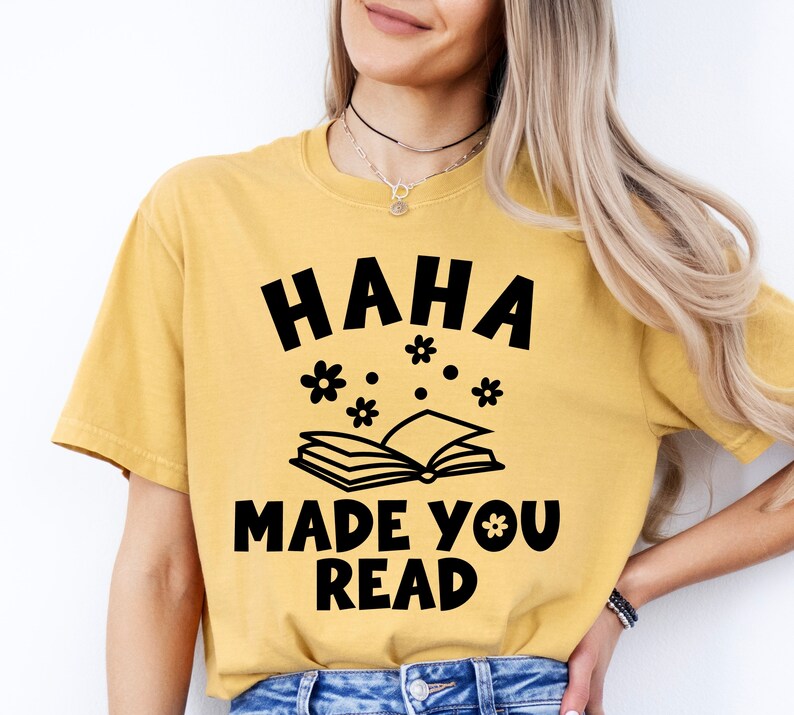 May include: A yellow t-shirt with black text that reads "HAHA MADE YOU READ" and a black illustration of a book with flowers above it.