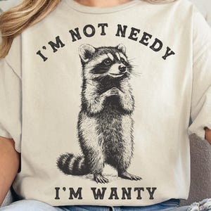 Puede incluir: Una camiseta beige con una ilustración en blanco y negro de un mapache. El texto "I'm not needy I'm wanty" está impreso en la camiseta.