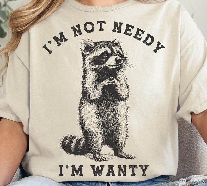 May include: A beige t-shirt with a black and white illustration of a raccoon. The text "I'm not needy I'm wanty" is printed on the shirt.