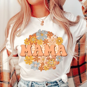 May include: A white t-shirt with a graphic design of the word "MAMA" in a retro font surrounded by colorful flowers. The flowers are in a circle and have a peach background.