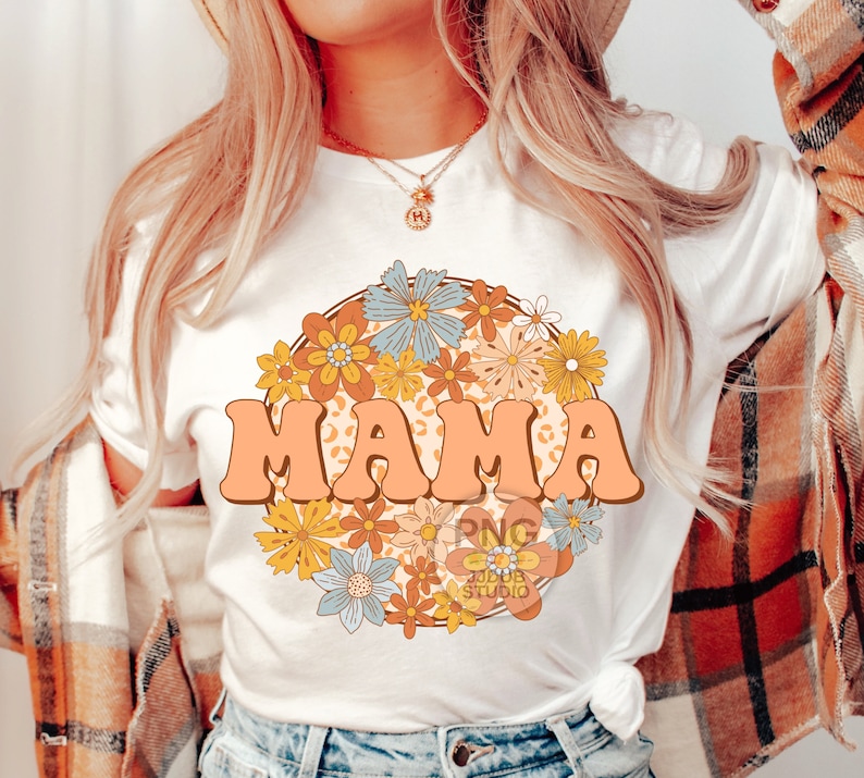 May include: A white t-shirt with a graphic design of the word "MAMA" in a retro font surrounded by colorful flowers. The flowers are in a circle and have a peach background.