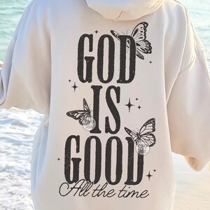 May include: Cream-coloured hoodie featuring a black graphic on the back. The design includes the words "GOD IS GOOD" with butterfly illustrations and the phrase "All the time". The hoodie has a hood and long sleeves.