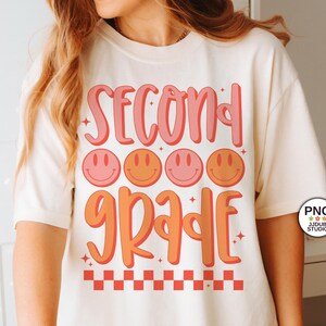 May include: A white t-shirt with the text "Second Grade" in pink and orange lettering. The text is surrounded by four orange and pink smiley faces. The bottom of the shirt has a red and white checkered pattern.