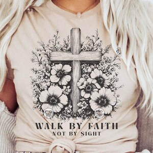 Peut inclure: Un t-shirt beige avec un motif noir et blanc d'une croix entour&eacute;e de fleurs. Le texte "Walk by Faith Not by Sight" est imprim&eacute; sous le motif.