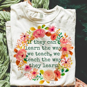 May include: Cream-colored t-shirt with a floral wreath design. The wreath features pink, orange, and yellow flowers with green leaves. The text reads: "If they can't learn the way we teach, we teach the way they learn."