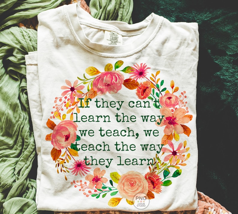 May include: Cream-colored t-shirt with a floral wreath design. The wreath features pink, orange, and yellow flowers with green leaves. The text reads: "If they can't learn the way we teach, we teach the way they learn."