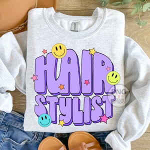 May include: A light gray sweatshirt with a purple and yellow graphic design that says "Hair Stylist" with smiley faces and stars.