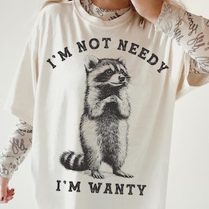 May include: A white t-shirt featuring a black and white illustration of a raccoon. The shirt has the text "I'm not needy I'm wanty" printed on it.