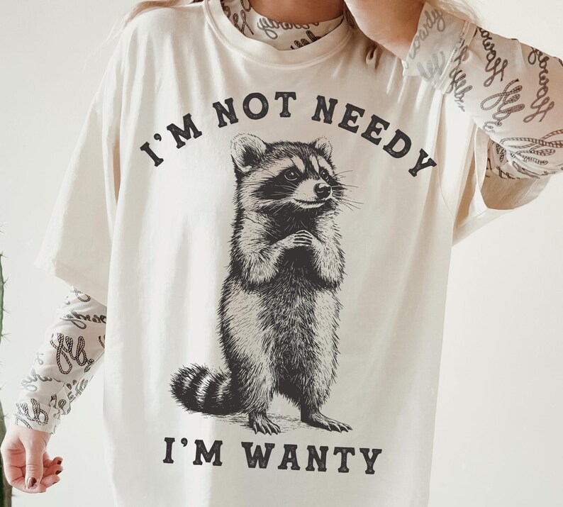 May include: A white t-shirt featuring a black and white illustration of a raccoon. The shirt has the text "I'm not needy I'm wanty" printed on it.