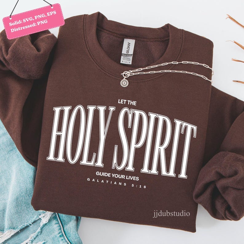 以下が含まれることがあります： 「LET THE HOLY SPIRIT GUIDE YOUR LIVES GALATIANS 5:16」の白い文字が入った茶色のスウェットシャツ。太陽のペンダントが付いたシルバーのネックレスがスウェットシャツの上にあります。スウェットシャツはジーンズの上にあります。