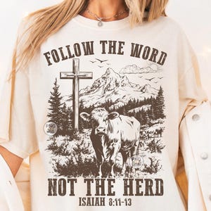 May include: A cream-coloured t-shirt with a graphic design. It features a cow, cross, and mountain landscape. The text reads "FOLLOW THE WORD" above and "NOT THE HERD" below, with "ISAIAH 8:11-13" at the bottom. The design is in shades of brown.