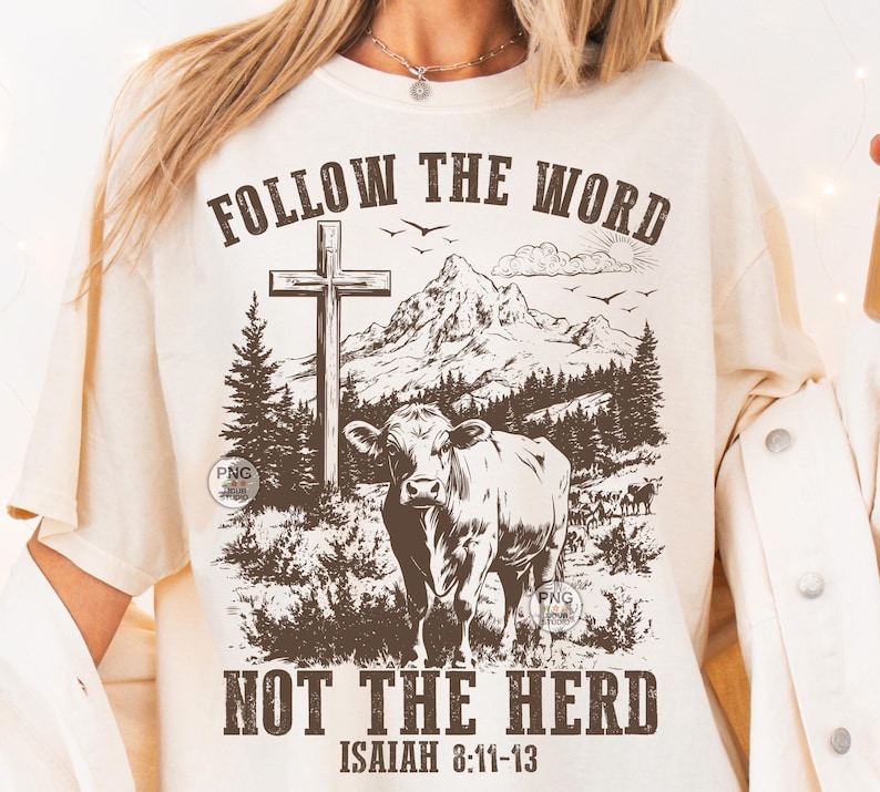 May include: A cream-coloured t-shirt with a graphic design. It features a cow, cross, and mountain landscape. The text reads "FOLLOW THE WORD" above and "NOT THE HERD" below, with "ISAIAH 8:11-13" at the bottom. The design is in shades of brown.