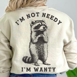 May include: A cream-coloured sweatshirt with a black and white raccoon graphic and the text "I'm not needy I'm wanty".