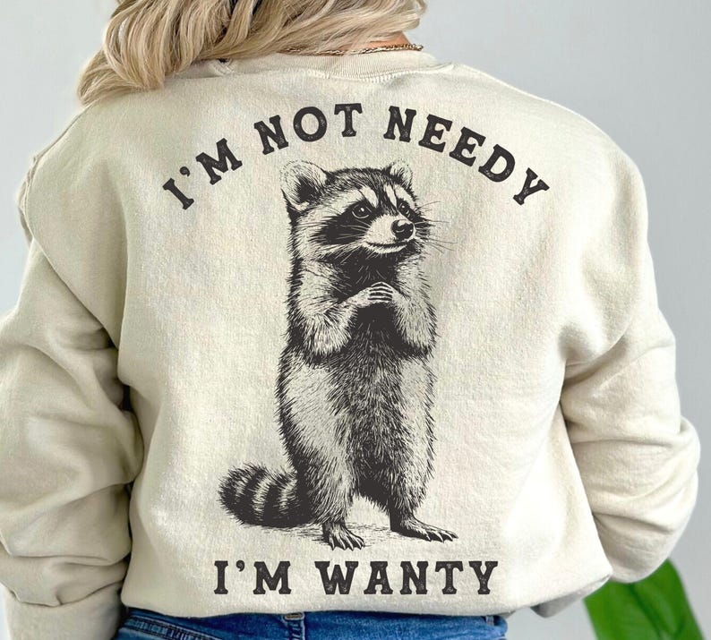 May include: A cream-coloured sweatshirt with a black and white raccoon graphic and the text "I'm not needy I'm wanty".