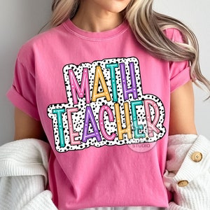 Op de afbeelding: Roze T-shirt met de tekst "MATH TEACHER" in een kleurrijk, retro-lettertype met zwarte stippen.