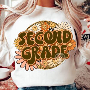 May include: White sweatshirt with a retro-style design featuring the words "Second Grade" in brown letters surrounded by colorful flowers and smiley faces.