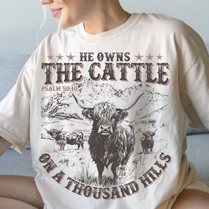 May include: A cream-coloured t-shirt featuring a Highland cow graphic and the text "HE OWNS THE CATTLE ON A THOUSAND HILLS" in brown. The design includes a mountain range and the Bible verse Psalm 50:10.