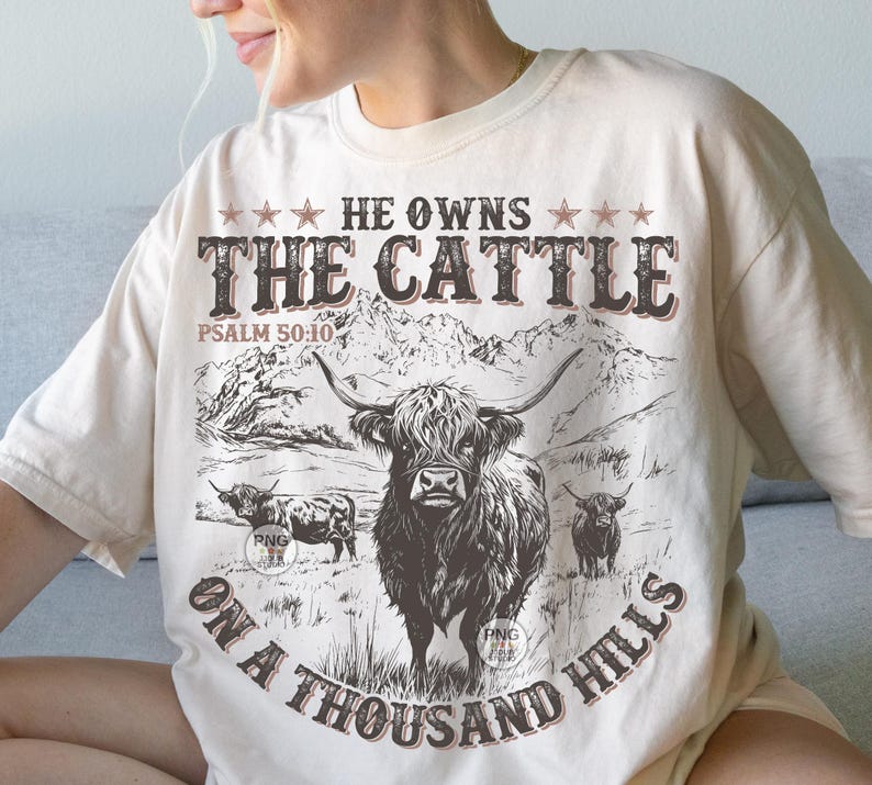 May include: A cream-coloured t-shirt featuring a Highland cow graphic and the text "HE OWNS THE CATTLE ON A THOUSAND HILLS" in brown. The design includes a mountain range and the Bible verse Psalm 50:10.